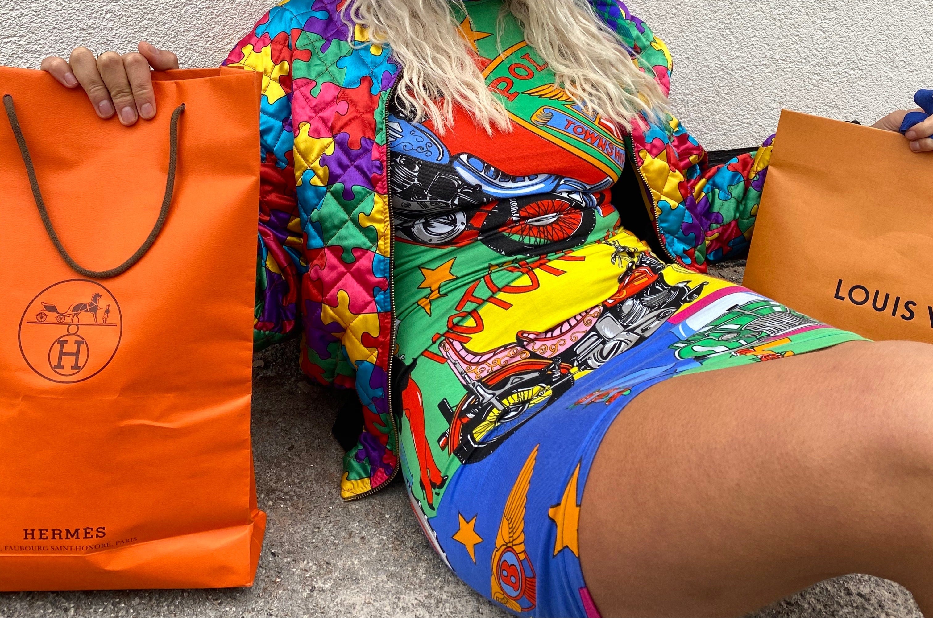 Person wearing a colorful outfit with a patterned headscarf, holding Hermes and Louis Vuitton bags.